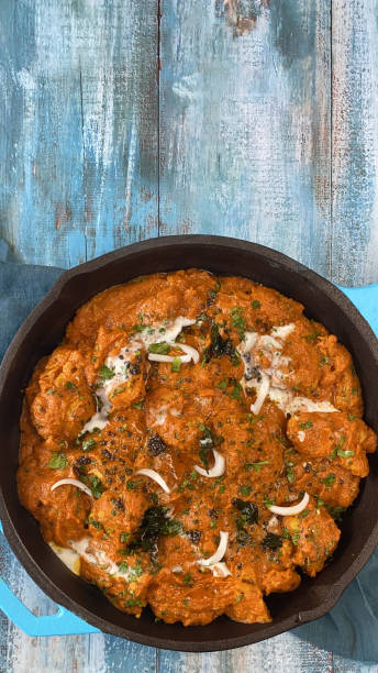 Stock photo showing elevated view of turquoise blue pan of chicken tikka curry sauce. Butter chicken breast chunks sprinkled with red onion and mustard seeds. imagen de una sartén azul turquesa llena de pollo con mantequilla / curry tikka, grandes trozos de carne de pechuga de pollo en salsa de curry con guarnición de rodajas de cebolla roja y semillas de mostaza, espacio de copia de fondo de grano de madera a