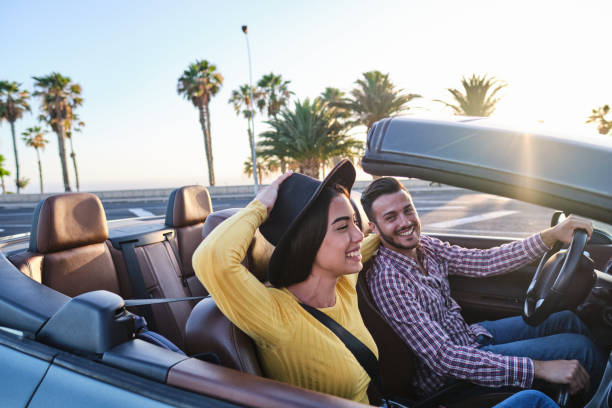 Young girl holding her hat while driving in a convertible car with her boyfriend Young girl holding her hat while driving in a convertible car with her boyfriend - Handsome young guy driving in the sunset with his beautiful girlfriend convertible stock pictures, royalty-free photos & images