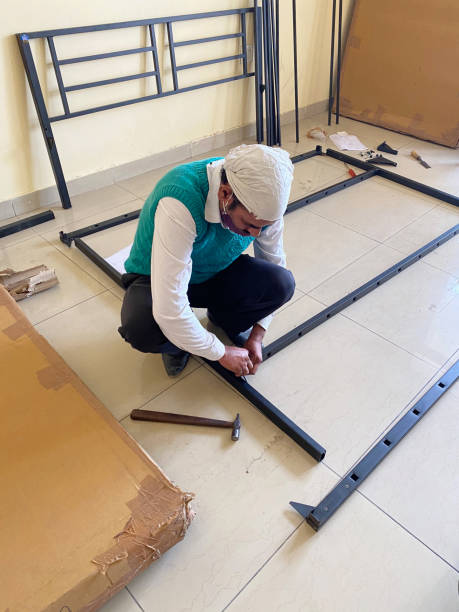 Image of Indian handyman crouching on tiled floor beside a cardboard box as he constructs some flat pack furniture, bed frame