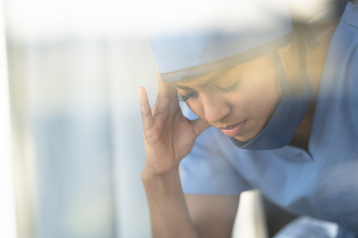 A close up of a female doctor appearing stressed and burnt out. She has her hand on her head as she is having a headache.