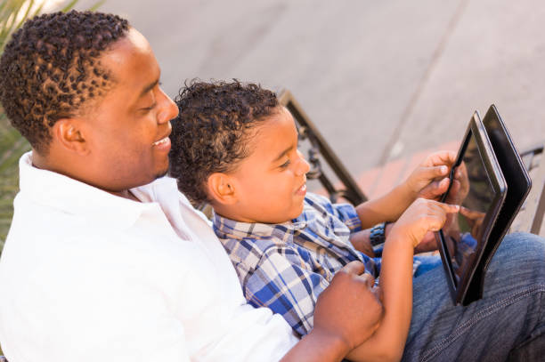 istock African American Father and Mixed Race Son Using Computer Tablet on Bench in Park 1301661116