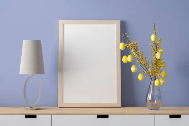 Sideboard with blank picture frame mockup, a desk lamp in easter egg form and easter eggs hanging on a forsythia twig in a glass vase. Easter eggs in illuminated yellow with different patterns.