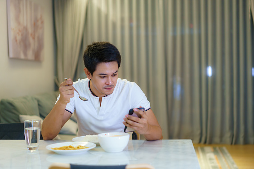 An Asian man is using his cell phone for chat with his friend or checking e-mail while eating dinner at home at night.