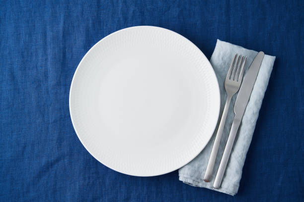 istock Clean empty white plate, fork and knife on bright blue dark linen tablecloth on table, copy space 1301689924