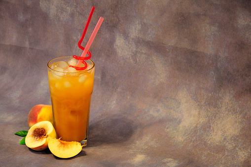 Tall glass of fruit nectar with ice and slices of ripe peach next to it on a gray background. View from above.