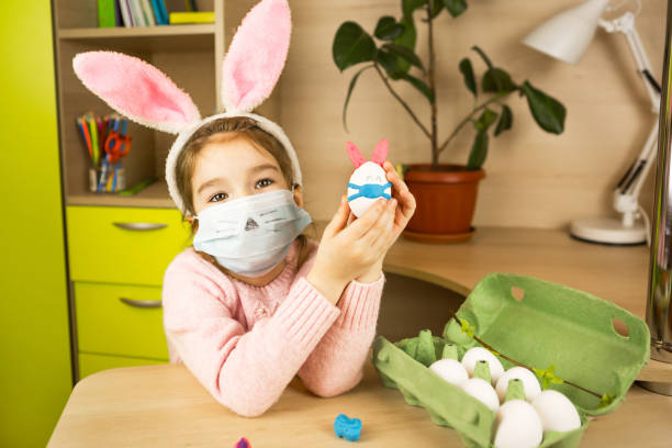 a girl in the ears of a hare makes an easter bunny in a medical mask out of an egg and plasticine. diy sitting at home, preparing for a religious holiday, home interior. - child easter egg home improvement easter imagens e fotografias de stock
