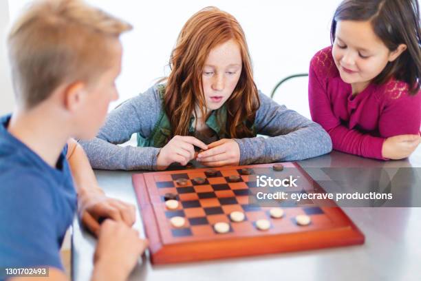 Three Siblings Playing Checkers Together At Dining Table Photo Series Stock Photo - Download Image Now