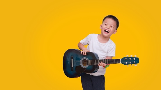 Asian happy smiling 5 years old boy having fun playing guitar isolated on colored background, Music for kids and toddlers concept