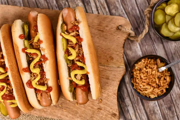 Homemade hotdogs with sausages and buns topped with preserved pickles, dried roast onions, mustard and ketchup next to bowls with ingredients