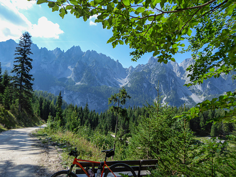 An orange mountain bike leaning against a bench next to a gravelled road in the mountains with the view on high Alps in the region of Gosau, Austria. The chains in the back are stony and barren. Calm