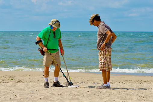 Cape May, New Jersey / USA - September 5, 2012: An older man uses a metal detector to hunt for buried objects at the beach as another man watches.