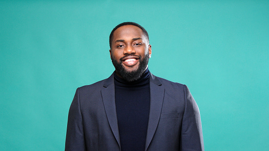 Sincere Afro American gentleman in turtleneck and jacket smiles showing white teeth touching chest by green studio wall closeup