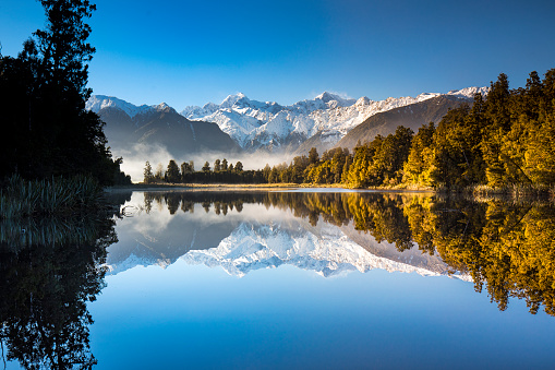 Golden snow capped mountain range reflecting perfectly on mirror lake with bright blue sky