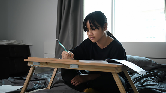 Photo of young Asian girl sitting on her bed and doing homework.