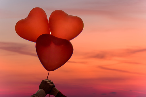 Valentines day. Silhouette of man and woman hands holding three reds balloons in form of heart on pastel sunset pink sky background. Beach. Concept