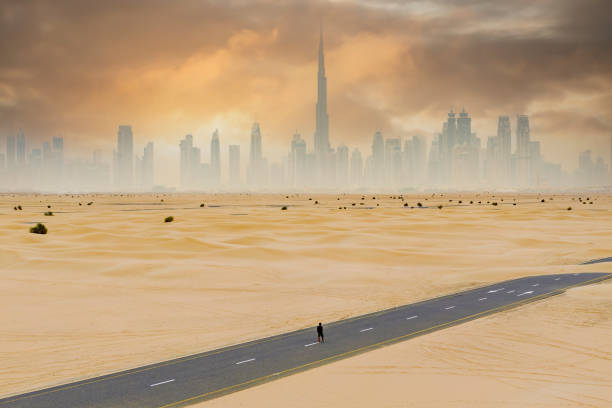 vista dall'alto, splendida vista aerea di una persona non identificata che cammina su una strada deserta coperta da dune di sabbia con lo skyline di dubai sullo sfondo. dubai, emirati arabi uniti. - fog desert arabia sunset foto e immagini stock