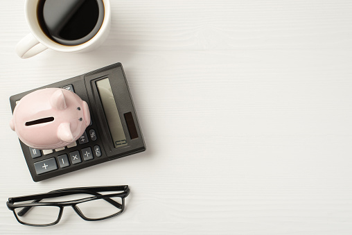 Flatlay top view photo of calculator with piggybank cup of espresso and eyeglasses isolated white wooden backdrop with empty space