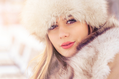 Beautiful woman with fur hat looking at camera in winter park in the city during the day in snowy weather with falling snow. Photo is shot with Sony a7III camera.