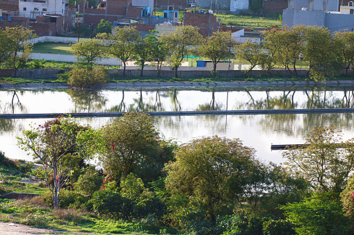 Stock photo of farming field and clean public sewage treatment plant in New Delhi suburbs, India, water being treated and purified for environment by filtration sewerage system, blue sky and trees making Indian sewage plant photo appear like lake wildlife, blue sky copy space