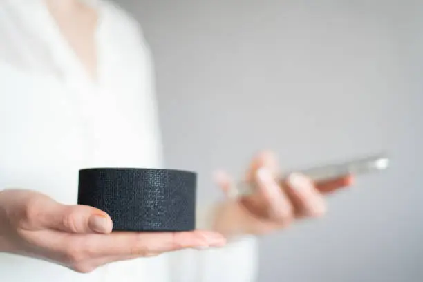 Photo of Synchronizing phone and a portable voice assistant speaker to control a smart home. Close-up of girl's hands hold for mobile phone to portable column