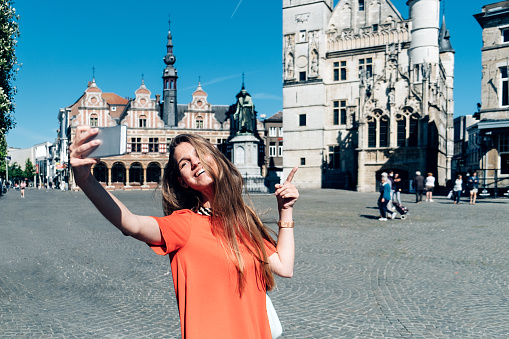 Pretty young woman taking a selfie in historic center of a belgian, european town - Beautiful girl walking on the streets as a tourist and taking selfie with landmarks