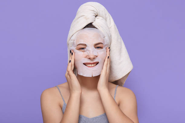 istock Adorable smiling lady wrapped in towel posing isolated over lilac background, using tissue mask for beauty procedures, keeping palms on cheeks, girl with bright manicure wearing sleeveless t shirt. 1301749679