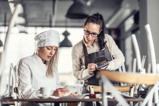 Female chef and waitress sit over bills calculating after hours in a restaurant. Female chef and waitress sit over bills calculating after hours in a restaurant. ipad calculator stock pictures, royalty-free photos & images