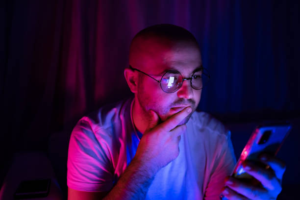 Young thoughtful man looking at his cellphone in blue and red neon light room stock photo