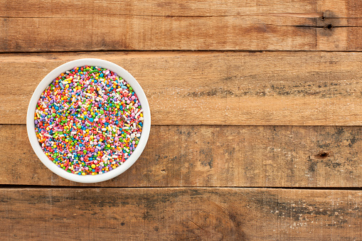 Top view of white bowl full of multi colored sprinkles over wooden table