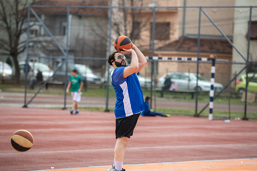 Overweight man with sports goggles practicing jump shot on outdoors basketball court