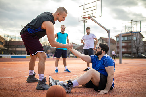 Young man helping his friend to get up on outdoor basketball court