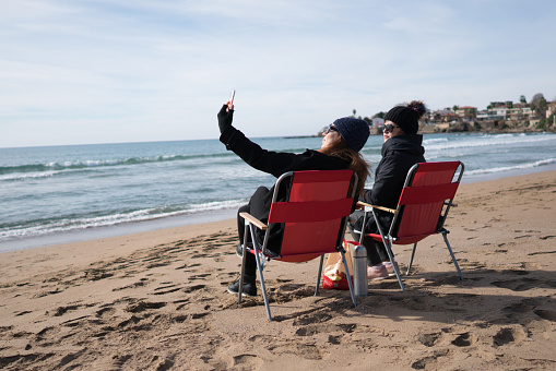 Two middle-aged women sit on the beach in winter and talk to each other.