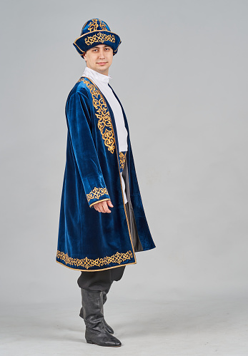 A young handsome aucasian man is dressed in a Kazakh folk clothing and a male headdress. The male dancer is standing half a turn to the camera looking at the camera. Studio shooting
