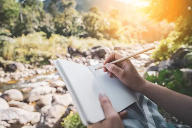 Photo of Woman hand writing on notebook with pencil at river background.