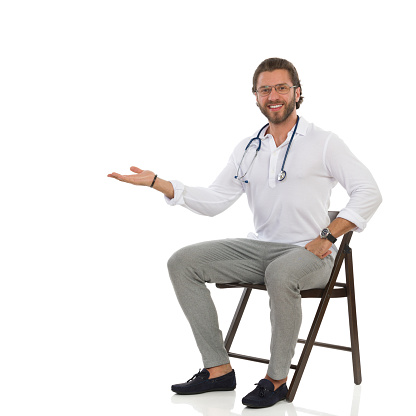 Handsome male doctor is sitting on a chair, holding hand raised, presenting something and smiling. Full length studio shot isolated on white.