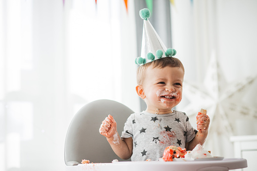 Baby boy celebrating first birthday in high chair and eating cake