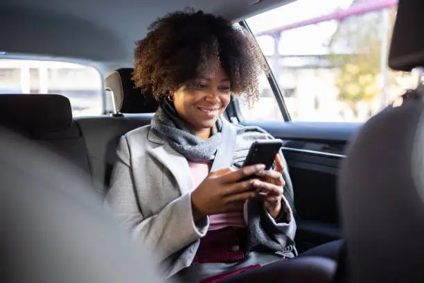 Woman sitting on backseat of a car and using mobile phone. African female traveling by a car.