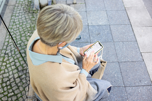 Overhead view of a senior woman wearing face mask sitting on a bench with using mobile phone at bus station. Senior commuter using mobile phone while waiting for bus in city during pandemic.