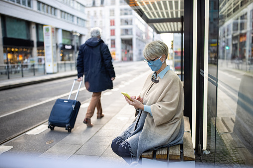 Senior woman wearing face mask sitting at bus station using mobile phone. Female commuter at tram station with a man walking by with luggage during pandemic.