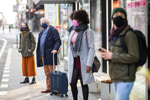 Diverse group of people wearing protective face masks standing at city transport station maintaining social distance. Men and women waiting at tram station.
