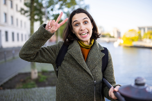 Portrait of a funny looking woman gesturing peace sign with her eyes wide open. Female tourist standing by a railing in the city posing for a photograph.