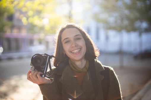 Beautiful woman smiling at camera. Female traveler holding her camera standing outdoors.