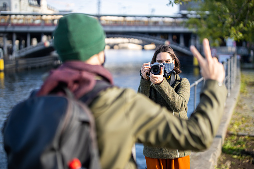 Woman taking photos of her boyfriend gesturing peace sign while on vacation. Female tourist wearing face mask filming her boyfriend on holiday.