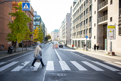 Side view of a senior woman wearing face mask crossing road using zebra crossing in the city. Mature woman traveling in the city during covid-19 outbreak.