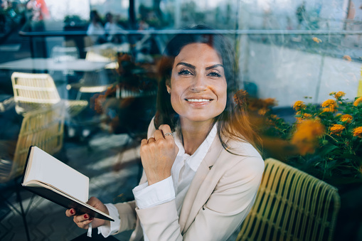Portrait of cheerful brunette business woman enjoying spending free time on hobby reading in cafe near window glass, smiling 30s intelligent female looking at camera holding notepad with planning