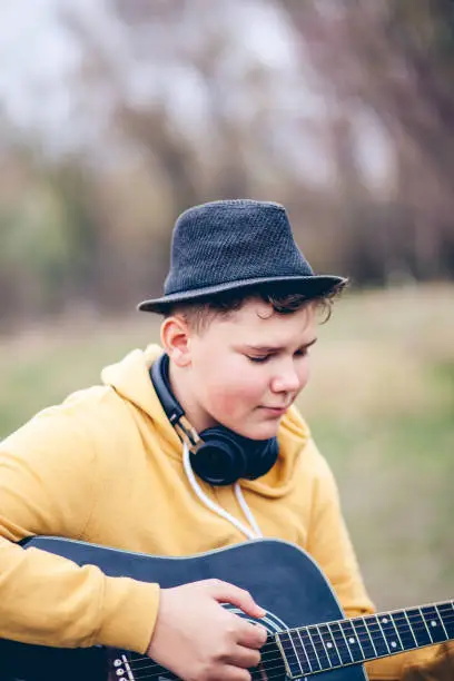 November 22, 2020 - Warsaw, Poland: close up portrait of a cute curly teenage boy playing on acoustic guitar, being involved, carried away, daydreaming while sitting in a meadow, wearing yellow jacket, fedora hat, eyes closed.