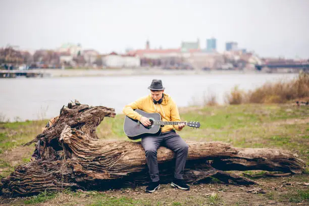 November 22, 2020 - Warsaw, Poland: focused, adamant adorable teen boy musician playing acoustic guitar on a riverbank in Warsaw with amazing scenic panorama behind, sitting on a tree trunk, wearing yellow hoodie, fedora hat.