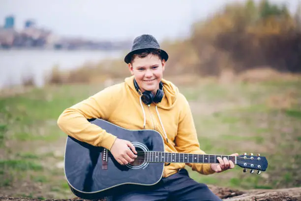 November 22, 2020 - Warsaw, Poland: Handsome intelligent cool teen boy playing guitar, smiling cheerfully, but shy, wearing headphones, fedora hat,  with a river behind.