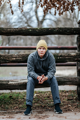 November 22, 2020 - Warsaw, Poland: handsome teen boy sitting on a wooden fence, hands clasped, fuul lenght, looking at camera seriously, confidently.