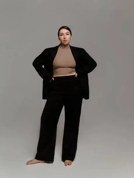 Successful business woman wearing black suit Blazer Pants and beige turtleneck Portrait of confident young lady Plus size fashion model studio portrait on gray background Full length body portrait looking at camera Barefoot